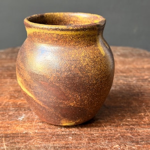 May include: A small, rounded ceramic vessel with a textured, mottled surface featuring earthy tones of deep brown, rust, and golden yellow. The pot has a wide, open mouth with a slightly flared rim and a smooth, rounded body shape.