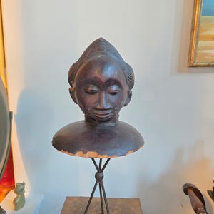 May include: A dark brown wooden bust sculpture of a person's head and shoulders. The sculpture has a stylized face with closed eyes and a small smile. The bust is mounted on a metal stand.