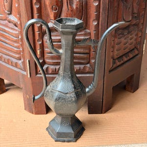 May include: A dark gray metal ewer with a hexagonal base, body, and neck. The vessel features an elongated curved spout, a tall handle, and intricate etched patterns covering the surface, including geometric shapes and stylized floral motifs.