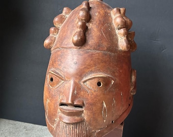 May include: A carved wooden sculpture of a human head with a beard and stylized facial features. The head is brown with a textured surface and is mounted on a reddish-brown base. The sculpture has a traditional, tribal aesthetic.