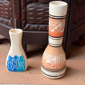 May include: Two ceramic vases of different heights. The shorter vase is cream-colored with a blue graphic design. The taller, slender vase features horizontal black bands and orange sections with white etched geometric patterns on a cream base.