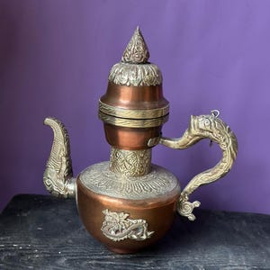 May include: An antique copper and brass teapot with ornate dragon and floral detailing. The teapot features a decorative handle and spout, and a conical lid. The body of the teapot is copper, while the handle, spout, and lid are brass.