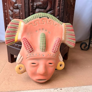 May include: A terracotta-colored ceramic wall mask featuring a stylized face with prominent ear spools, three vertical corn-shaped ornaments on the forehead, and a green-striped headdress with red and yellow accents and geometric carvings.