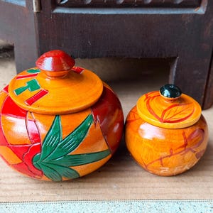 May include: Two round wooden lidded containers with a glossy finish. The larger container features a vibrant orange, red, and green leaf pattern. The smaller container has a yellow and orange base with a dark knob and subtle painted leaf details.