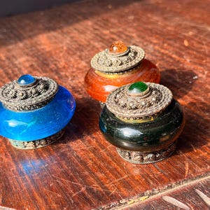 May include: Three small, round containers with translucent glass bodies in blue, amber, and dark green. Each container features a decorative silver-toned metal lid with intricate floral patterns and a small, smooth, colored cabochon stone at the center.