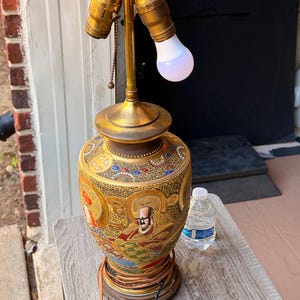 May include: A table lamp with a brass fixture and two bulb sockets, one containing a white LED bulb. The base is a ceramic vase featuring intricate, multi-colored hand-painted figures, gold accents, and detailed patterns against a dark background.