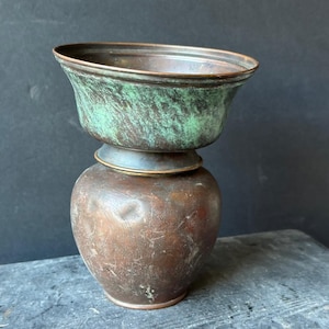 May include: An antique copper vessel with a two-part design. The top section is a bowl with a green patina, while the base is a rounded pot with a brown finish. The vessel sits on a gray surface against a dark background.