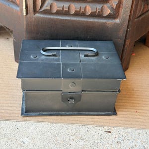 May include: A rectangular black metal storage box featuring a flat lid with a centered handle, a front latch mechanism, and visible rivet details on the top surface. The box has a matte finish and a structured, boxy design with clean edges.