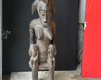 May include: A carved wooden sculpture of a seated figure with a detailed face and headdress. The figure has prominent breasts and holds two smaller figures on its lap. The sculpture is a light brown color and appears to be aged.