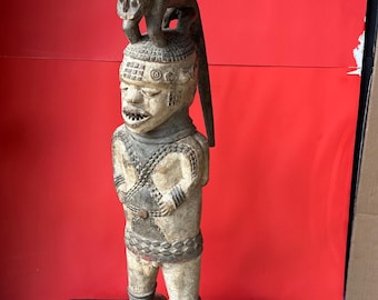 May include: A carved wooden statue of a figure with a monkey on its head. The statue is painted in shades of beige and brown, with intricate details on the clothing and face. The figure is standing on a black base against a red background.