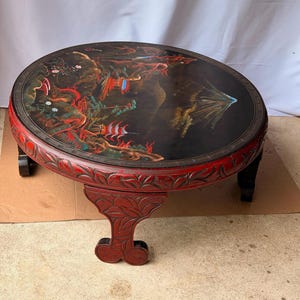 May include: A round, low-profile coffee table with a black lacquered top featuring a painted landscape with a mountain, trees, and structures. The table has a deep red, carved wooden frame and legs with intricate leaf-like patterns.