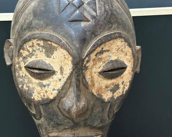 May include: A carved wooden mask with a dark brown finish. The mask features a prominent forehead with a cross-like design, closed eyes, and a detailed mouth. The eye sockets have a lighter, weathered appearance. The mask is mounted on a black stand.