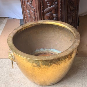 May include: A large, round, hammered brass planter with a wide, flat rim and two decorative lion head handles on the sides. The interior shows a natural patina with hints of green oxidation at the base, contrasting with the polished gold exterior.