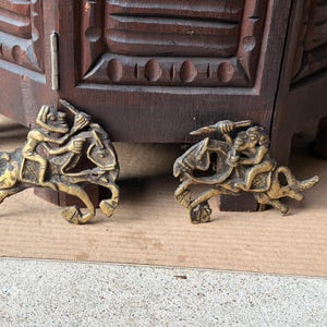 May include: Two identical brass-colored metal ornaments featuring a figure riding a horse. Each piece depicts a rider holding a spear or staff while mounted on a galloping horse. The metal has a dark, aged patina with highlights on the raised surfaces.