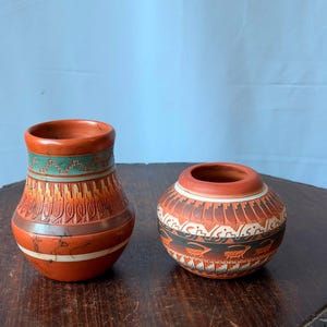 May include: Two terracotta-colored ceramic vessels featuring intricate hand-painted geometric patterns and animal motifs. The taller vase has a teal band near the rim, while the shorter, rounded pot displays a repeating pattern of horned animals.