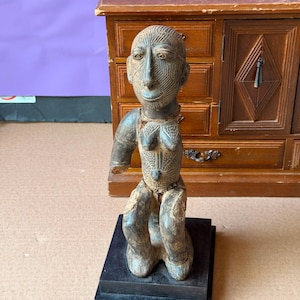 May include: A dark brown carved wooden figure in a seated position, featuring intricate linear patterns etched across the face, chest, and torso. The figure has a missing right arm and is mounted on a square black base, displaying a weathered texture.