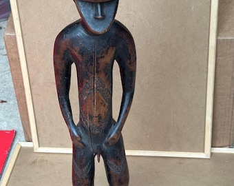 May include: A carved wooden figure of a standing person with a stylized face and body. The figure is dark brown with carved details on the torso and legs. The figure is approximately 12 inches tall.