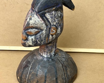 May include: A carved wooden sculpture of a stylized human head. The head has a dark blue headdress and a face with white and brown tones. The sculpture is mounted on a rounded base, showcasing a rustic, aged appearance.
