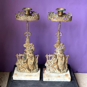 May include: Pair of antique brass candle holders. Each holder features a detailed design with a central stem, a decorative base with figures, and a circular top for the candle. The bases are set on square marble blocks.
