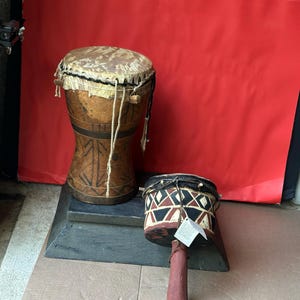 May include: Two handcrafted drums. The larger drum is brown with a patterned design and a natural hide drumhead. The smaller drum has a geometric pattern in black, red, and white, with a red handle. Both drums are on a dark gray surface.