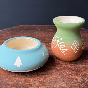 May include: A matte turquoise ceramic bowl with a white tree graphic and a small matte green and terracotta ceramic vase featuring white geometric line patterns. Both vessels have a smooth, cream-colored interior finish and rounded shapes.