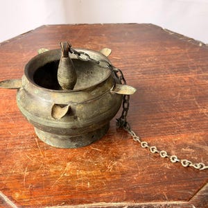 May include: A weathered bronze hanging vessel featuring a rounded bowl with a wide rim, four small protruding tabs, and a central vertical handle. A metal chain is attached to the handle and extends outward, showing signs of oxidation and age.