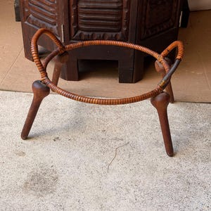 May include: A circular stand featuring a brown wooden frame with three tapered legs and a rim wrapped in thin, coiled rattan material. The structure includes two arched handles extending upward from the main ring, providing a stable base.
