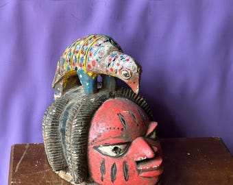 May include: A carved wooden mask with a red face, black hair, and a colorful bird figure on top. The mask has white eyes and black markings. The bird is painted with blue, yellow, red, and white spots. The mask is on a wooden surface.