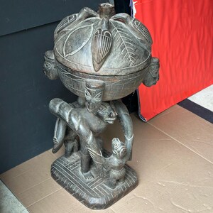 May include: A carved wooden sculpture featuring a spherical top with leaf and face details, supported by figures. The sculpture has a dark brown finish and rests on a rectangular base. The piece appears to be a decorative or functional item.