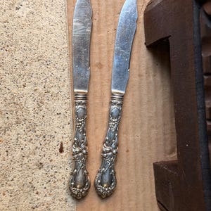 May include: Two silver-toned metal knives featuring ornate, embossed floral and scrollwork patterns on the handles. The blades are smooth, slightly curved, and show signs of surface wear. The handles have a detailed, raised relief design.