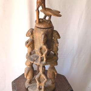 May include: A tall, brown, hand-carved wooden sculpture featuring a central cylindrical container topped with a bird figure. The structure is supported by two tiers of carved human figures standing on circular wooden platforms, showcasing intricate details.