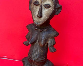 May include: A carved wooden figurine with a stylized human form. The figure has a light-colored face and dark brown body, with prominent breasts and a detailed abdomen. The figure is posed with bent legs and arms, set against a red background.