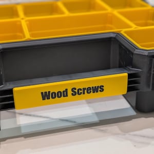 May include: A black and yellow storage container with multiple compartments. The container has a yellow label that reads "Wood Screws" in black text. The container is designed for organizing small hardware items.