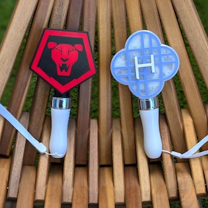May include: Two light sticks with white handles and lanyards. One has a red and black shield with a lion design. The other has a light blue, four-leaf clover design with the letter "H".