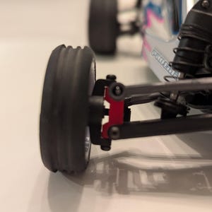 May include: Close-up of a black rubber tire on a small, remote-controlled car. The tire is mounted on a black suspension arm with a red component. The car's body is visible in the background, with white, blue, and pink accents.