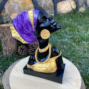 Könnte beinhalten: Eine schwarze Büste einer Frau, die einen lila und goldenen Kopftuch, goldenen Schmuck und ein gold-schwarzes Kleid trägt.