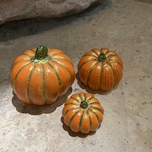 May include: Three decorative pumpkins in various sizes. The pumpkins are orange with gold and green accents. The largest pumpkin is in the center of the image, with a smaller pumpkin to the left and the smallest pumpkin to the right.