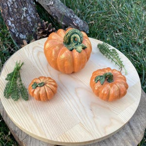 May include: Three orange pumpkins of varying sizes on a light wood surface. The largest pumpkin has a gold sheen and a green stem. The smallest pumpkin is in the foreground.