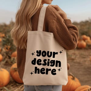 Könnte beinhalten: Beige Tragetasche mit dem Text "your design here" in Schwarz, akzentuiert mit Sternformen. Die Tasche hat cremefarbene Griffe und wird vor einem Kürbisfeld gehalten.