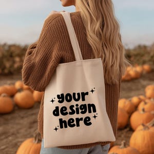 Könnte beinhalten: Cremefarbene Tragetasche mit dem schwarzen Text "your design here". Die Tasche hat eine rechteckige Form und lange Träger. Der Hintergrund zeigt Kürbisse und eine Person in einem braunen Pullover.
