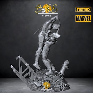 Può includere: Una statuetta in 3D stampata in grigio di due personaggi Marvel, Daredevil e Wolverine, che combattono. La statuetta è posizionata su una base di cemento rotta con un idrante e una scala. Il testo "TESTED" e "MARVEL" è visibile sullo sfondo.