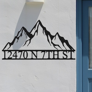 Peut inclure: Panneau d'adresse en métal noir avec une silhouette de montagnes et l'adresse "12470 N 7TH ST". Le panneau est fixé sur un mur blanc, à côté d'une porte bleue.