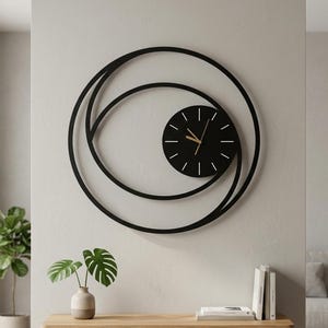 Puede incluir: Un gran reloj de pared circular con un marco de metal negro compuesto por dos anillos superpuestos. Una esfera de reloj circular negra más pequeña está colocada descentrada dentro del marco, mostrando marcadores de hora blancos y manecillas doradas.