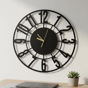 Puede incluir: Un reloj de pared redondo con un diseño de marco abierto en color negro y grandes números negros en negrita del uno al doce. El centro es un círculo negro sólido con manecillas finas de color dorado para horas, minutos y segundos.