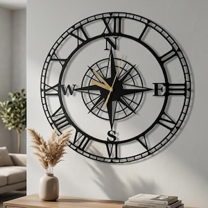 Könnte beinhalten: Eine große, runde Wanduhr mit einem schwarzen Metallrahmen, römischen Ziffern und einem zentralen Kompassrosen-Design. Die Uhr verfügt über dünne, goldfarbene Zeiger für Stunden, Minuten und Sekunden. Die Himmelsrichtungen N, O, S und W sind in der Mitte angebracht.