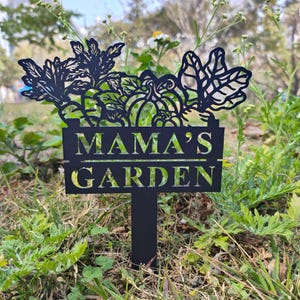 Puede incluir: Estaca de jardín de metal negro con un letrero que dice "Mama's Garden". El letrero está decorado con un diseño recortado de verduras y calabazas.