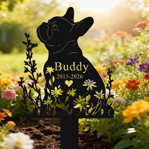 May include: Black metal garden stake featuring a silhouette of a French Bulldog. The dog's name, "Buddy", and years "2015-2026" are displayed in gold lettering. The stake is decorated with floral cutouts and is placed in a garden.