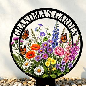 May include: Black metal garden sign with the words "GRANDMA'S GARDEN" in a circular frame. The sign features colorful flowers, butterflies, and a variety of blooms, including poppies, daisies, and asters. The sign is placed on a bed of small stones.