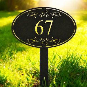 May include: Black oval address sign with the number 67 in yellow. The sign has a gold border and decorative flourishes. The sign is mounted on a black stake and is set in a grassy yard.