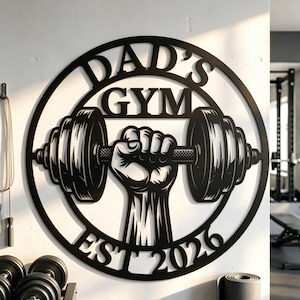 Könnte beinhalten: Runde Wanddekoration aus schwarzem Metall mit der Aufschrift "DAD'S GYM" und "EST 2026". Das Design zeigt eine Hand, die eine Langhantel hält. Ideal für ein Fitnessstudio oder einen Fitnessbereich zu Hause.